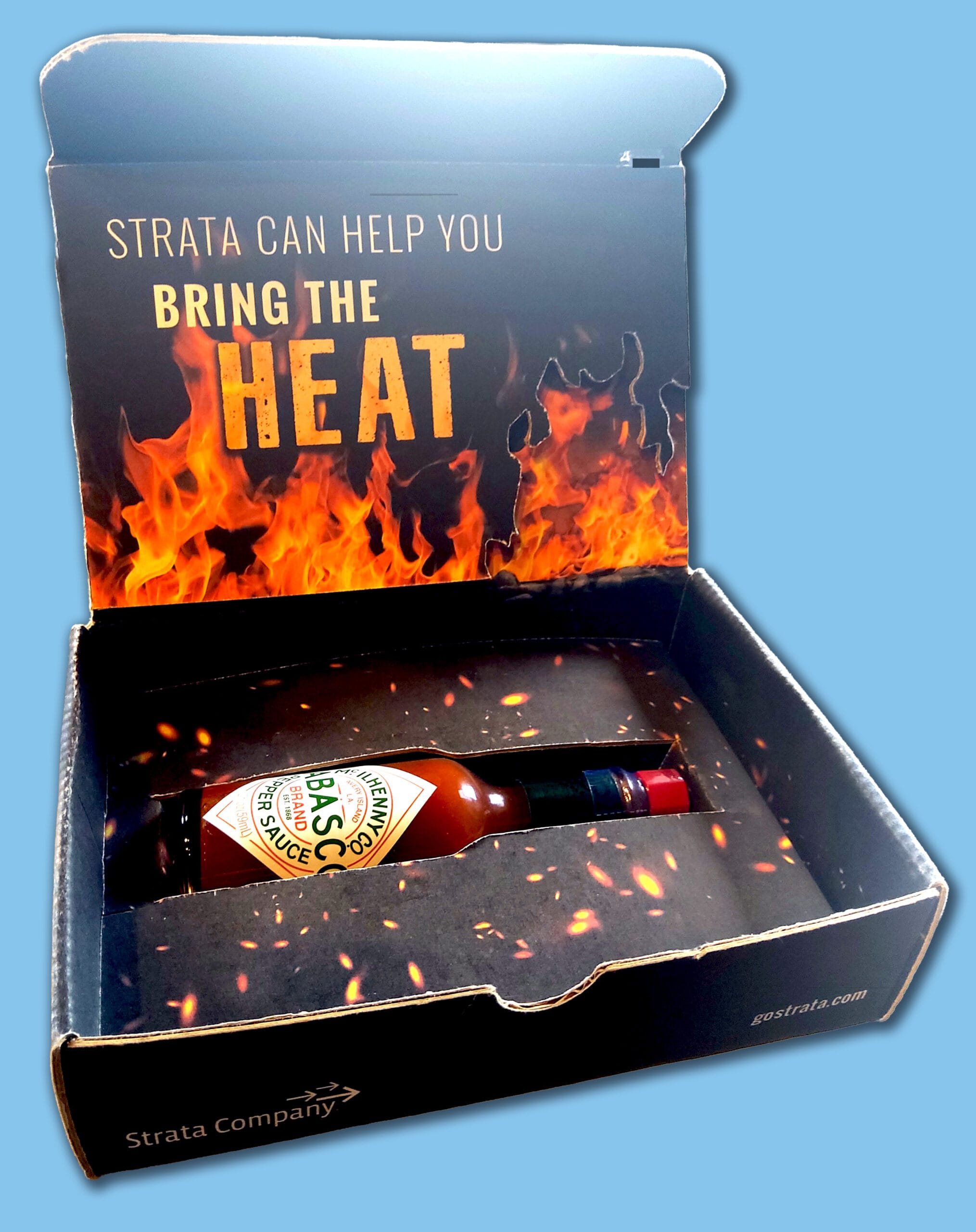 strata-box-insert-closed-blue-background Open promotional box from Strata Company containing a bottle of Tabasco pepper sauce. The inside lid features flames and the text: 'Strata can help you bring the heat.' The box interior has a fire and ember design, with the bottle secured in a custom cutout. Strata Company logo and gostrata.com are printed on the box sides.