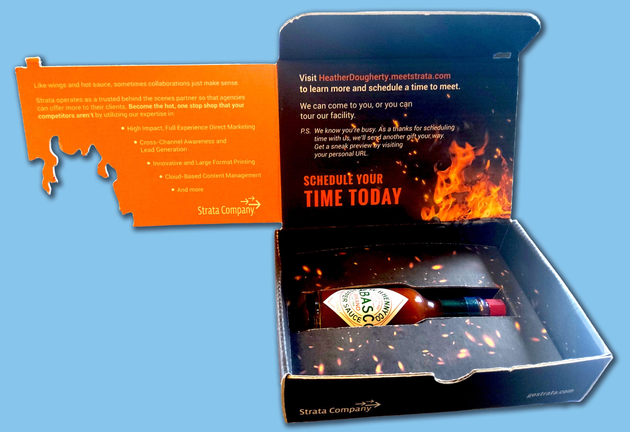 strata-box-insert-open-blue-background Open Strata Company promotional box containing a bottle of Tabasco pepper sauce. The left inside flap has an orange insert with marketing text about agency collaboration and Strata’s services. The right inside lid shows flames and text inviting recipients to schedule a meeting with 'Schedule Your Time Today' in bold orange letters. The interior bottom has a fire and ember design with a custom cutout holding the hot sauce bottle.