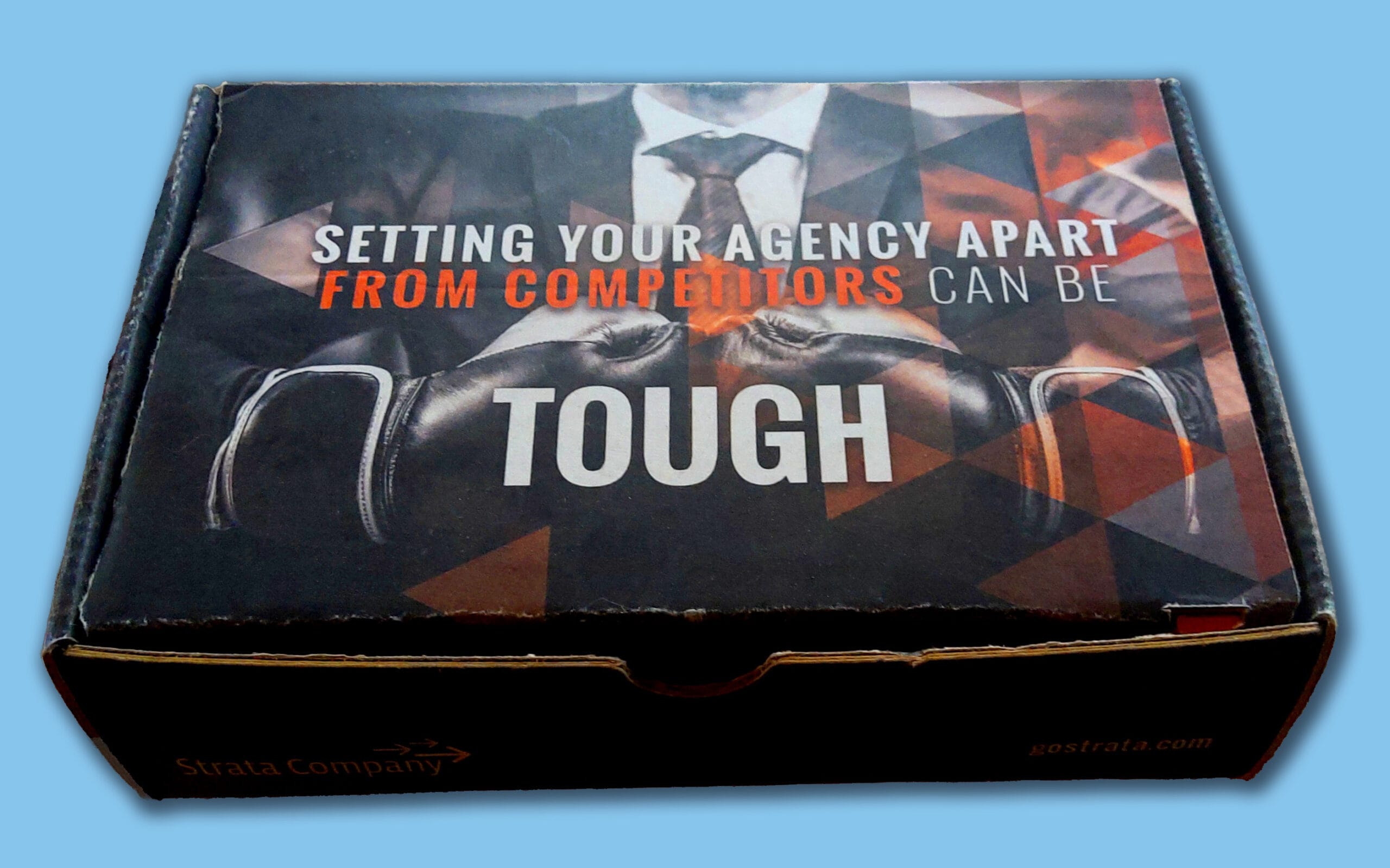Strata Hot Sauce_Exterior_LtBlue Rectangular black box with a design showing a man in a suit and tie wearing black boxing gloves. The text reads: 'Setting your agency apart from competitors can be tough.' The words 'from competitors' are in red, and the rest are in white. The bottom edge has the Strata Company logo and website address